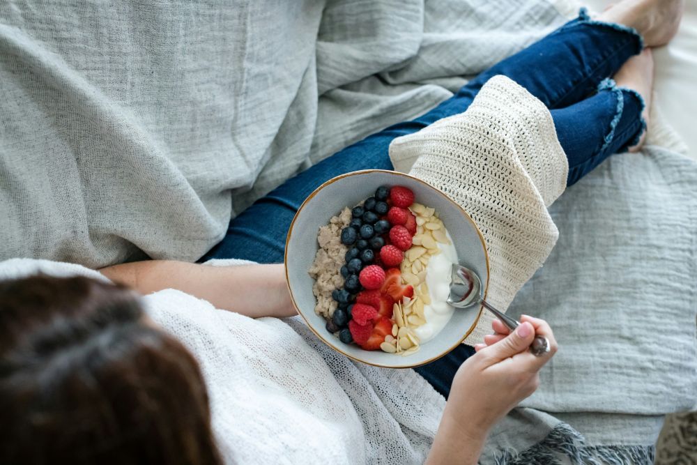 A woman relaxing on a couch enjoying a nourishing bowl of yogurt and fruit, representing a peaceful relationship with food and emotional eating recovery in Sydney.